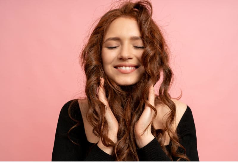 Mulher ruiva com cabelo cacheado e sorrindo de olhos fechados. Representando a satisfação após fazer cachos com chapinha de forma correta